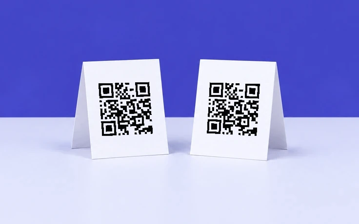 Deux chevalets blancs portant chacun un QR code noir aux modules nets, posés sur une table lavande pâle devant un mur indigo.